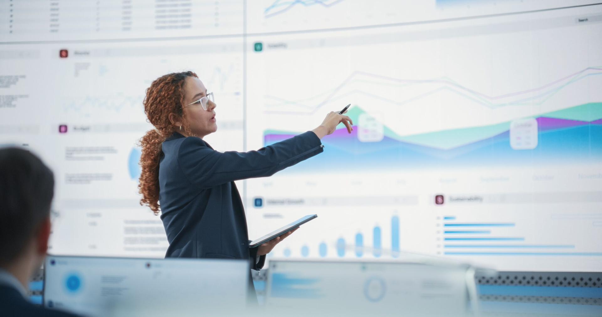 A woman in a blazer presents data visualizations on a wall in front of desktop computers
