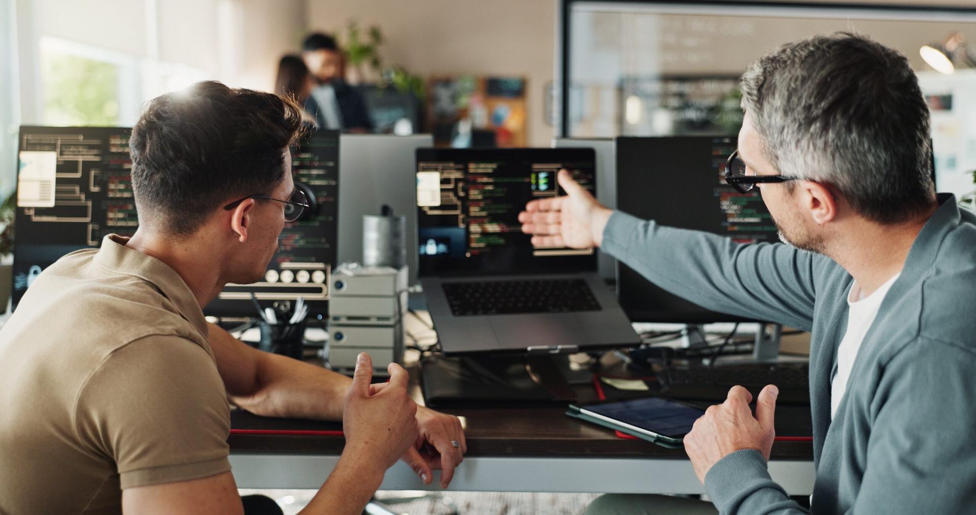 A man gestures to a computer monitor while a younger man looks on.