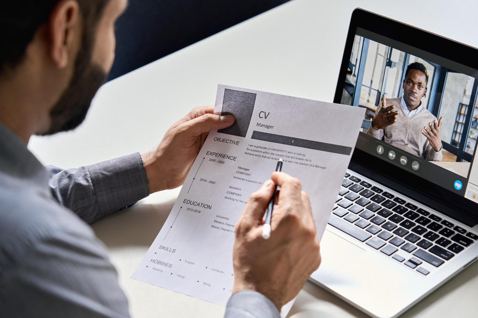 A man looks at a resume with a pencil while interviewing a candidate over Zoom, with the laptop open and the candidate visible on the screen